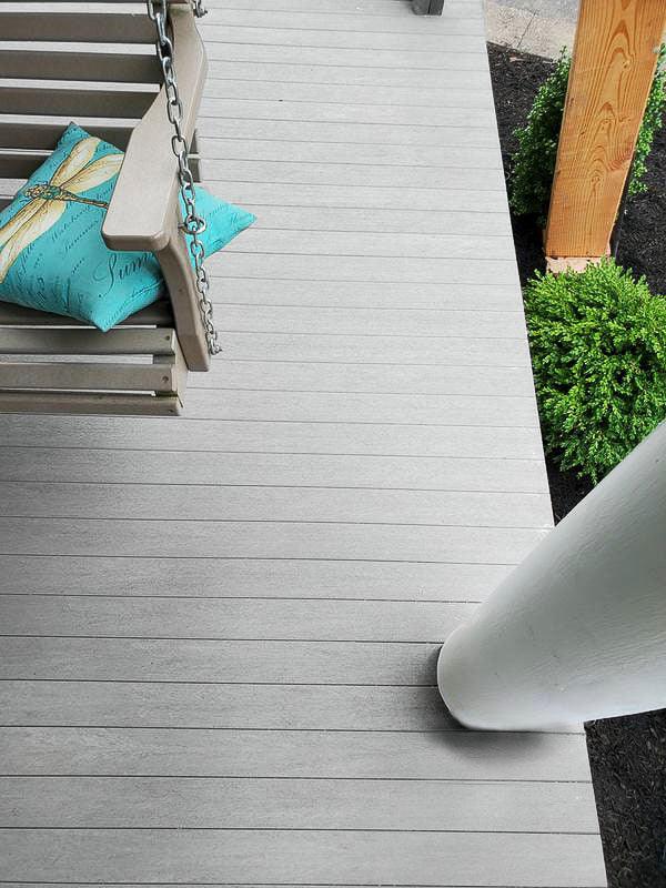 Terrasse en bois composite gris clair avec balancelle et coussin décoratif, poteau blanc et arbustes