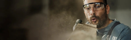 Homme soufflant la poussière sur une planche de bois, lunettes de protection, ébénisterie.